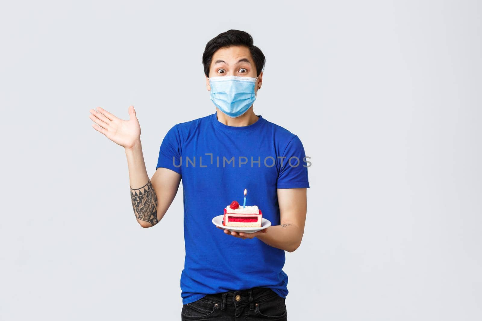 Self-quarantine, home lifestyle and celebration concept. Excited young man celebrating birthday, asian guy in medical mask waving hand and holding b-day cake, staring astonished.