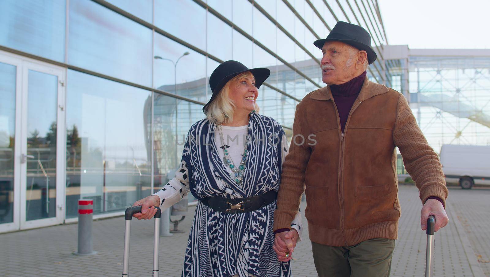 Elderly old husband and wife retirees tourists go to airport terminal for boarding or railway station for traveling. Lovely mature couple grandmother grandfather carrying luggage suitcases on wheels