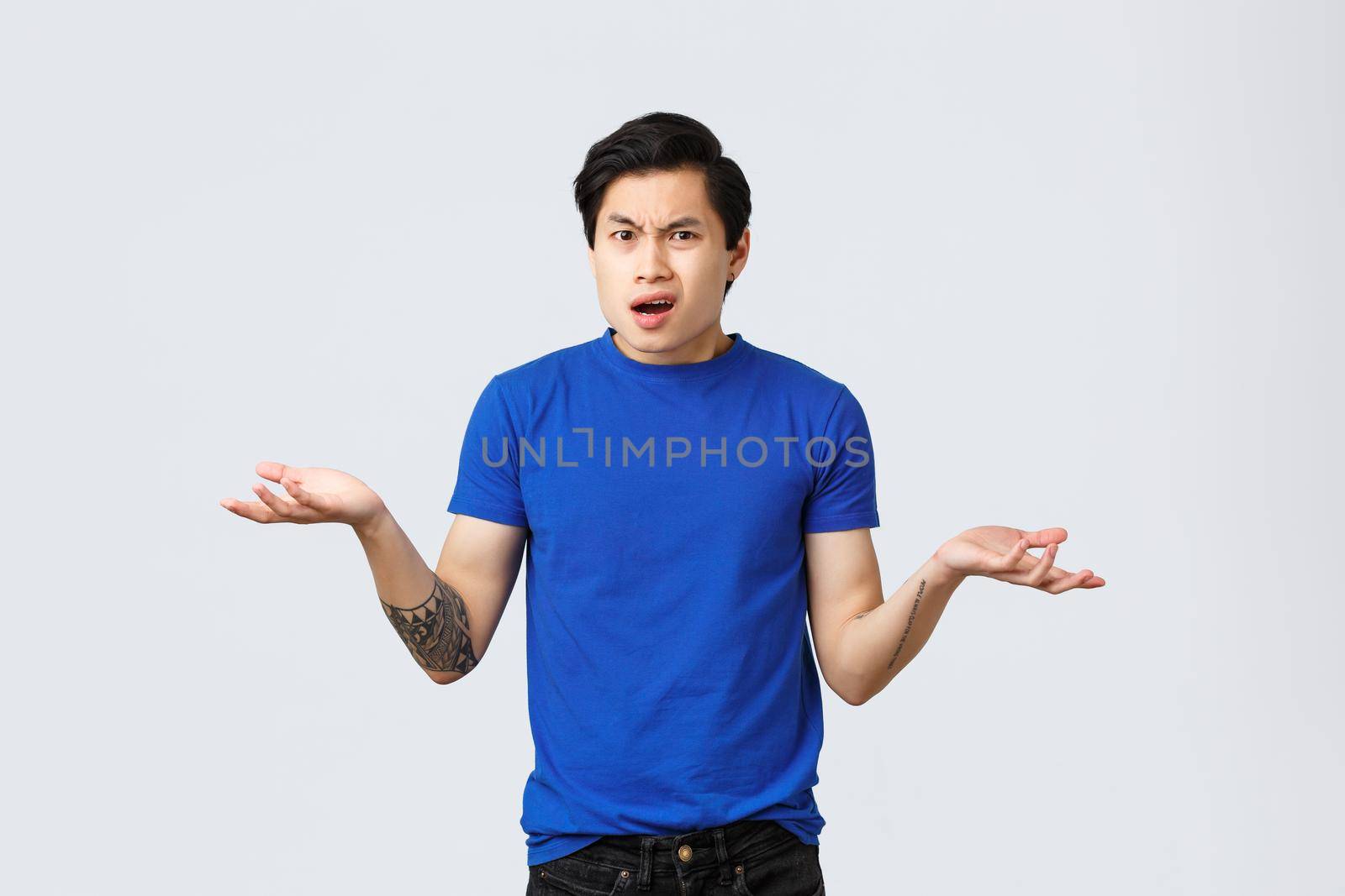 What is that. Confused and pissed-off young male employee, asian guy in blue t-shirt, shrugging with hands spread sideways, being frustrated, cant understand what happened, stand grey background.
