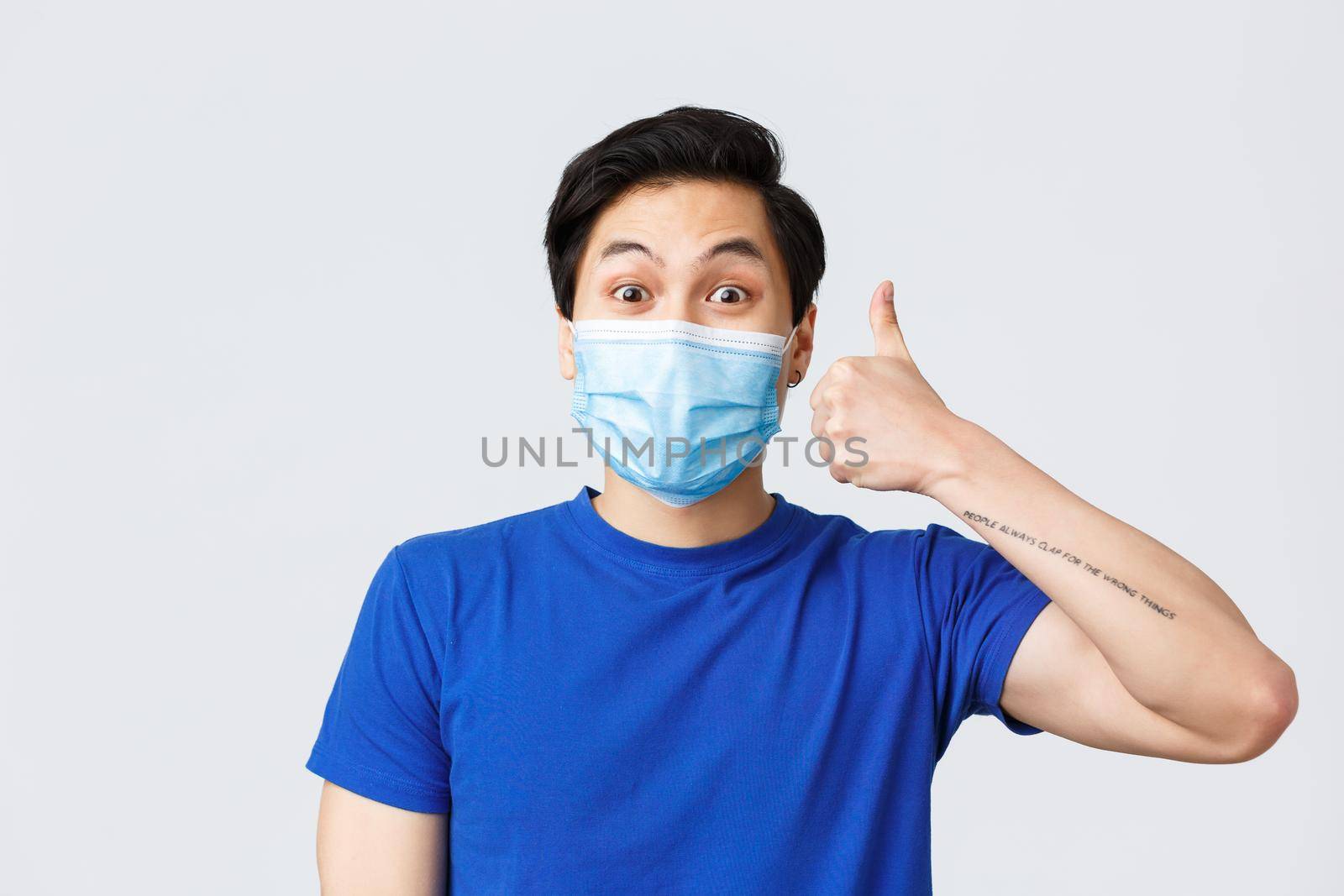 Different emotions, lifestyle and leisure during coronavirus, covid-19 concept. Enthusiastic happy asian man in medical mask and blue t-shirt show support and approval, make thumb-up.