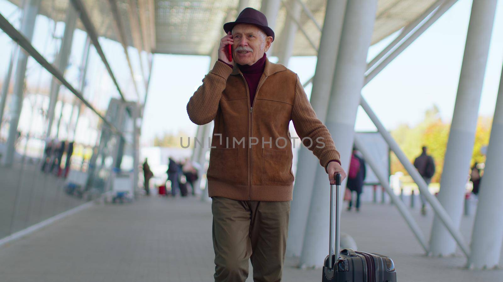 Senior tourist grandfather walking on international airport hall, railway station carrying luggage suitcase talking on smartphone and smiles. Travel, vacation. Pensioner businessman using mobile phone