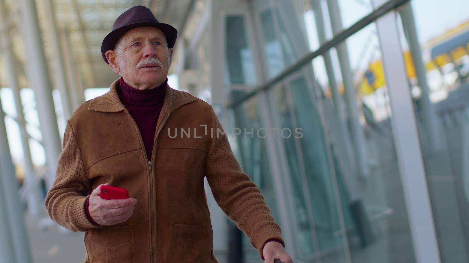 Senior tourist grandfather walking on international airport hall carrying luggage suitcase texting Messages on Smartphone and smiles. Travel, vacation. Pensioner businessman using mobile phone