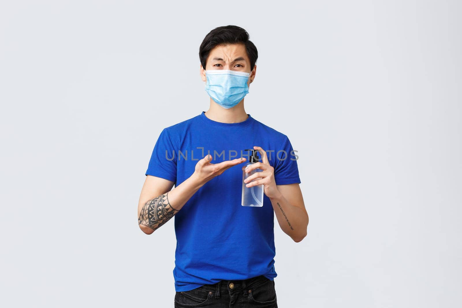 Lifestyle, people different emotions and covid-19 pandemic concept. Disgusted and upset grimacing asian man in medical mask hurry up to use hand sanitizer after handshake, grey background.