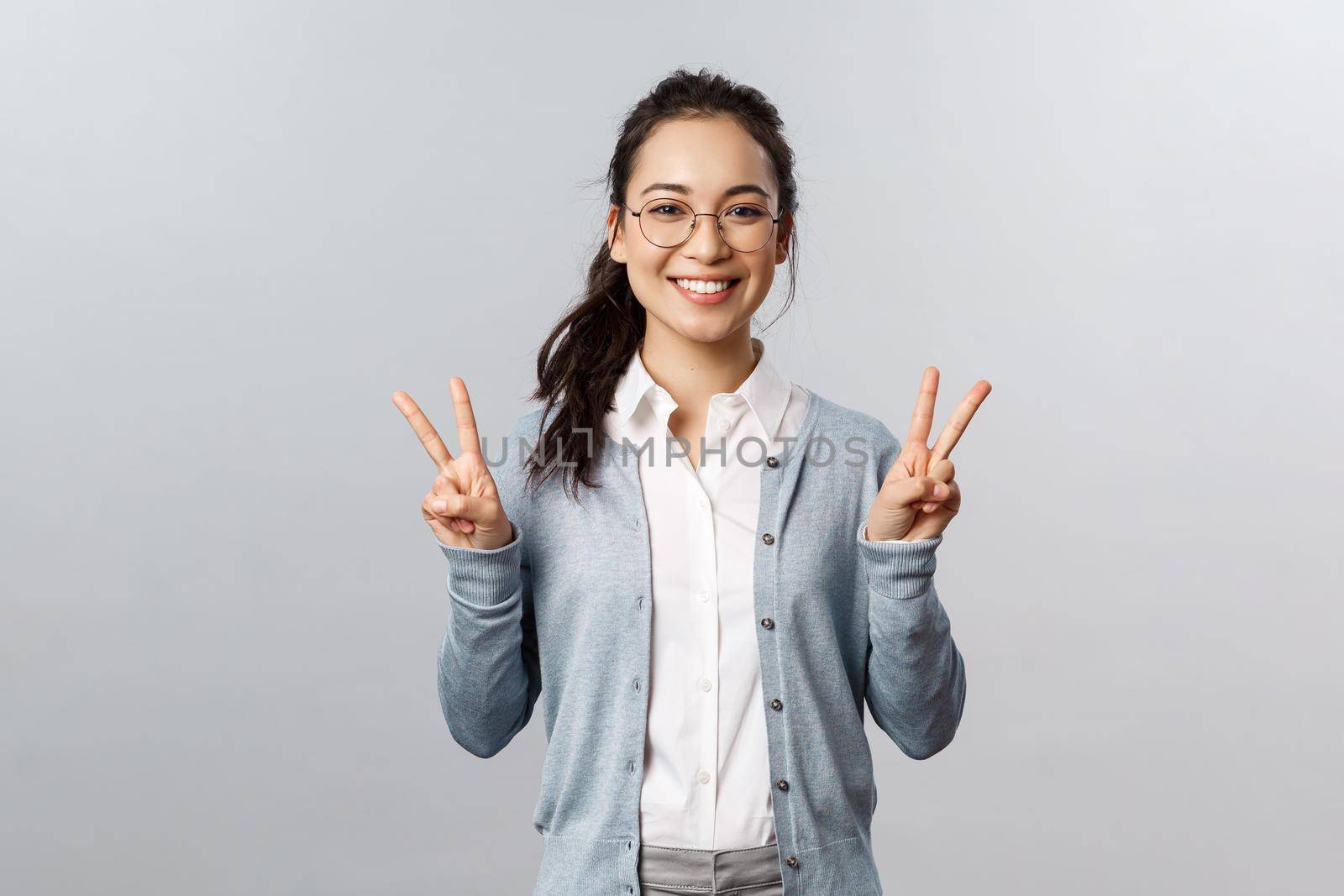 People, emotions and lifestyle concept. Staying on bright side. Cheerful smiling asian girl in glasses, showing peace signs kawaii, standing grey background friendly and rejoicing.