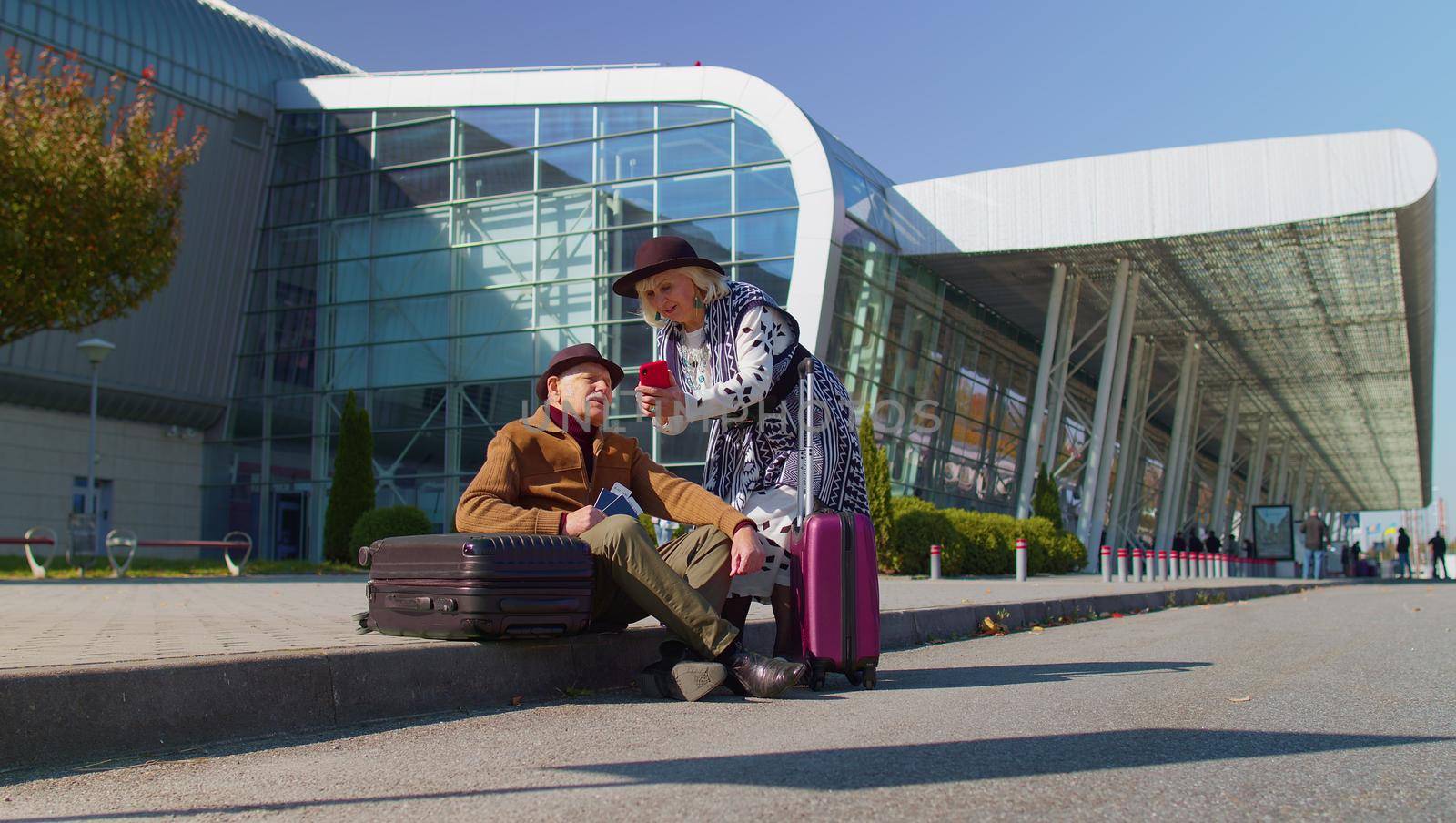Senior couple happy pensioner tourists grandmother grandfather waiting for boarding near international airport hall or railway station with luggage suitcases on wheels. Travel, vacation, journey, trip