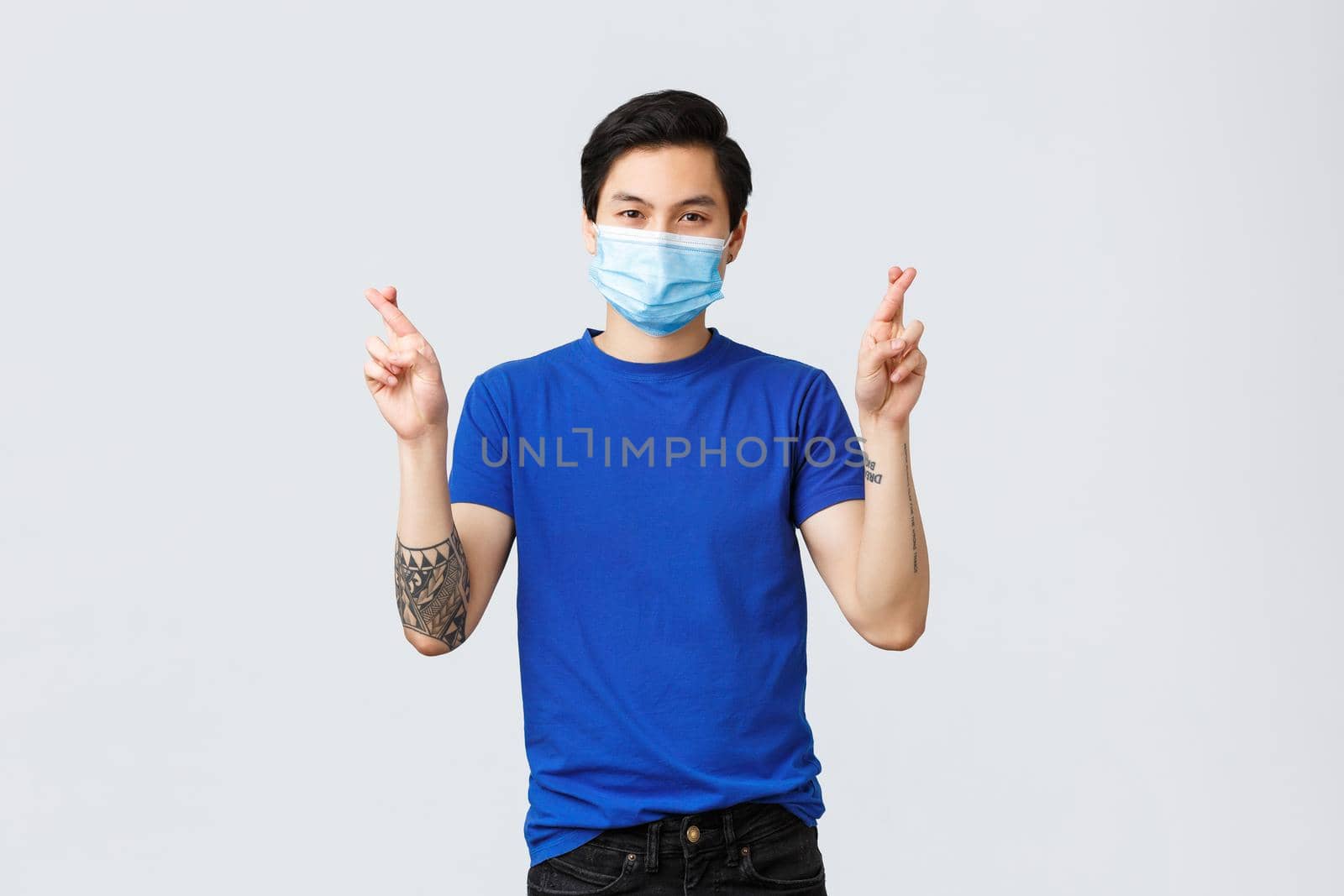 Different emotions, social distancing, self-quarantine on covid-19 and lifestyle concept. Determined hopeful asian man praying, cross fingers good luck, wearing medical mask.