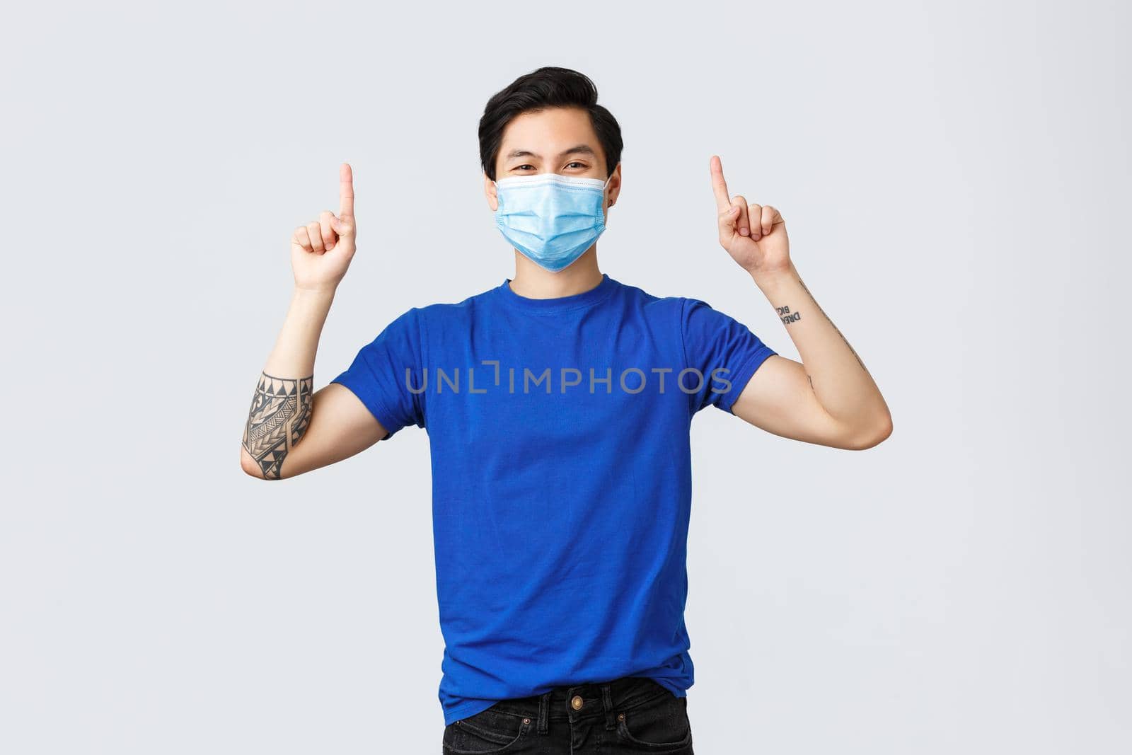 Different emotions, social distancing, self-quarantine on coronavirus and lifestyle concept. Cheerful smiling asian man in medical mask and t-shirt, pointing fingers up to advertise, showing banner.