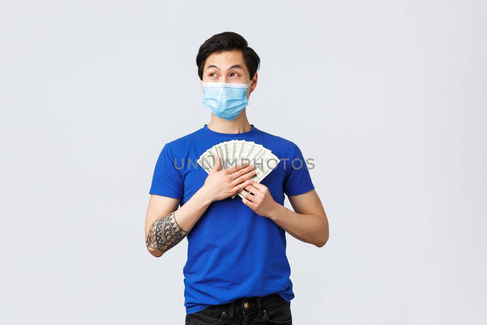 Money, lifestyle, insurance and investment concept. Dreamy happy asian man holding cash and thinking what buy on it, looking away smiling, wear medical mask, grey background.