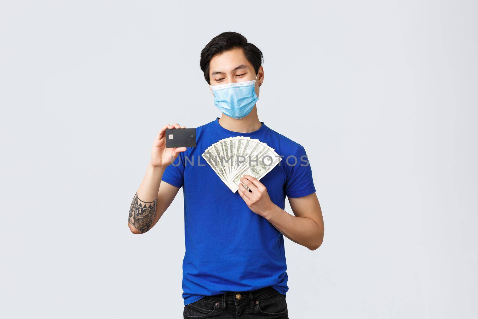 Money, covid-19, easy payment, investment and banking concept. Pleased young asian man in medical mask likes protect his income and health, place cash on deposit, smiling at credit card.