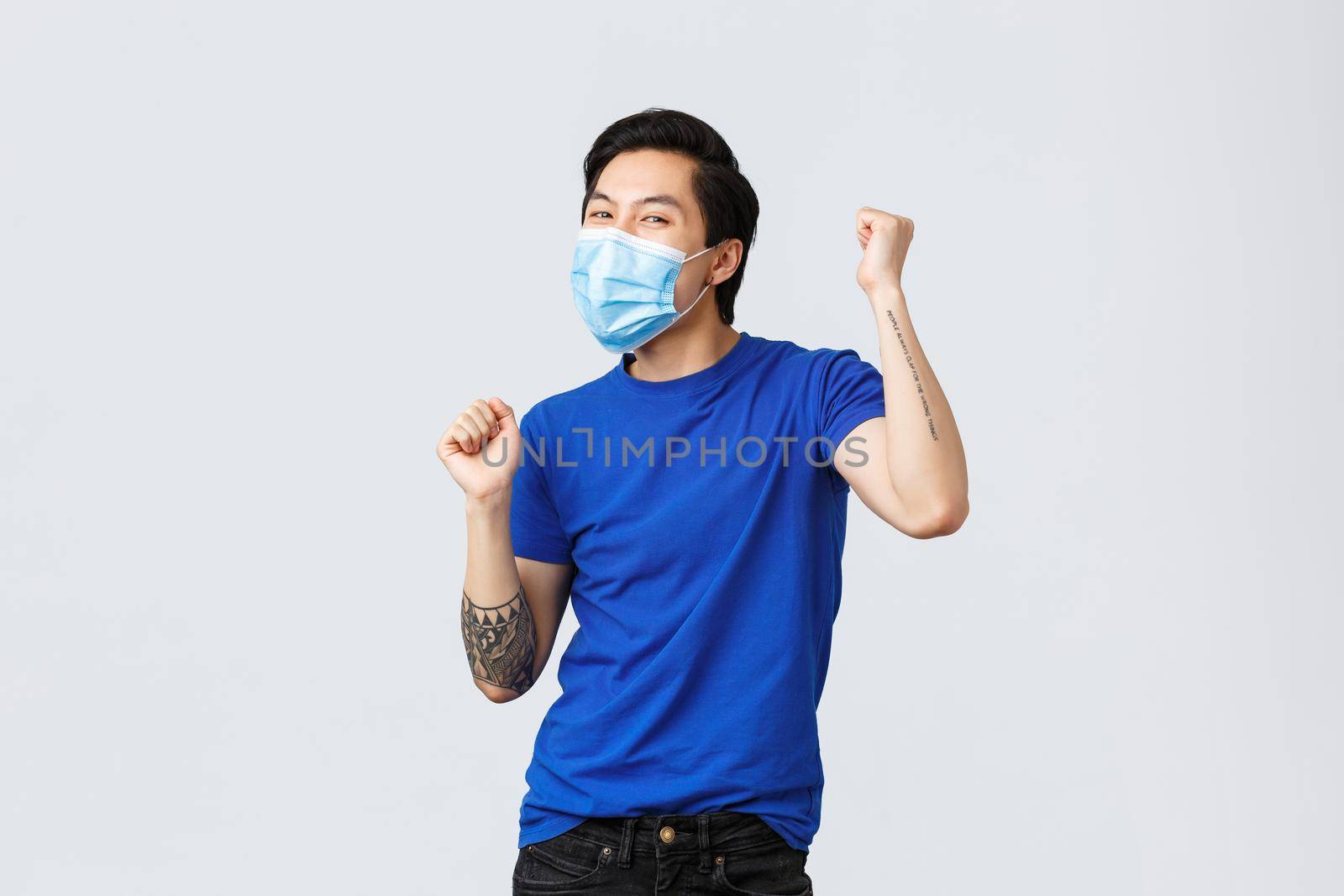Different emotions, social distancing, self-quarantine on covid-19 and lifestyle concept. Cheerful, carefree asian guy in medical mask, chanting, dancing from happiness, winning, achieve success.
