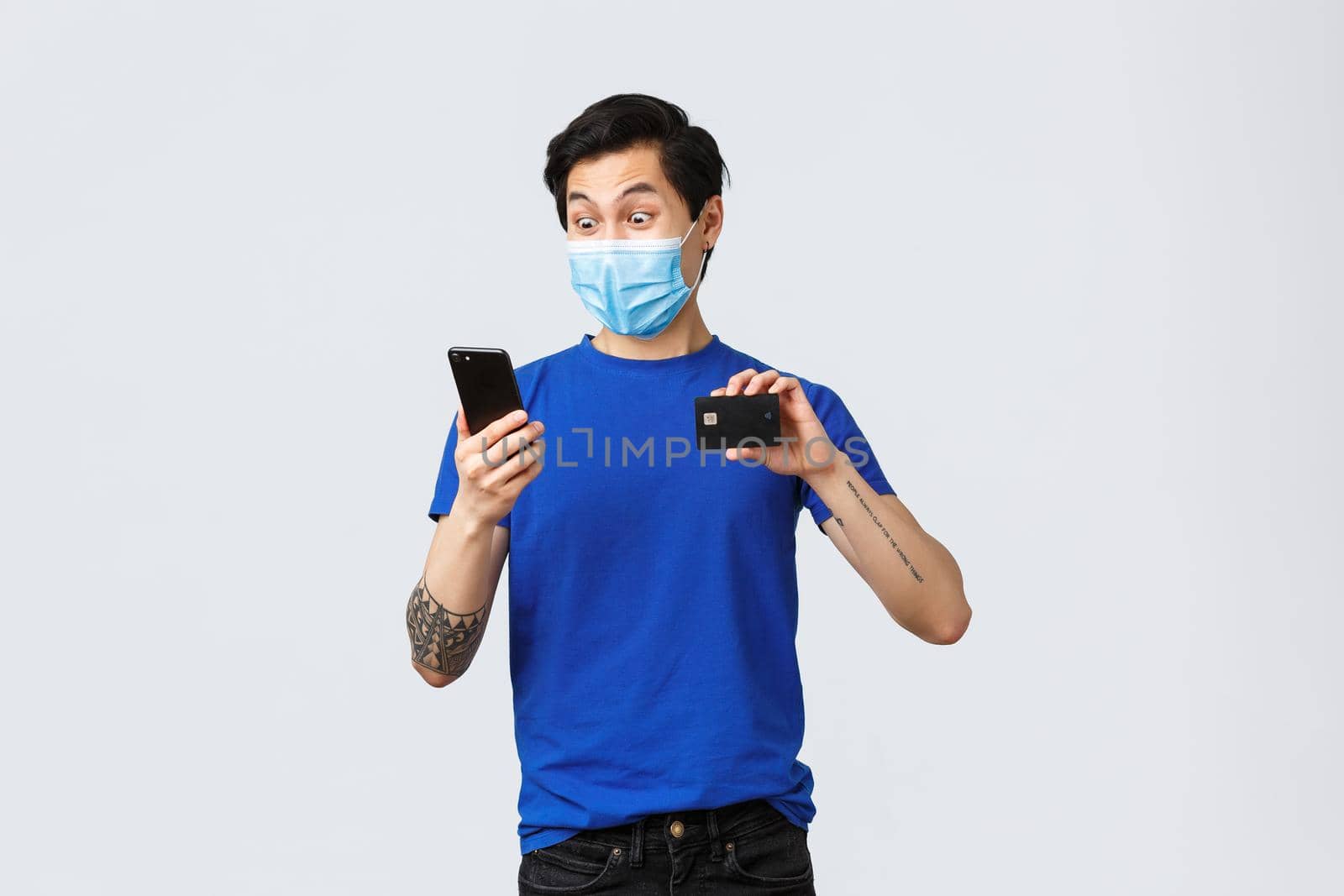 Contactless payment, online shopping during covid-19 and pandemic concept. Excited and amazed asian guy in medical mask staring astonished at smartphone banking account app, show credit card by Benzoix