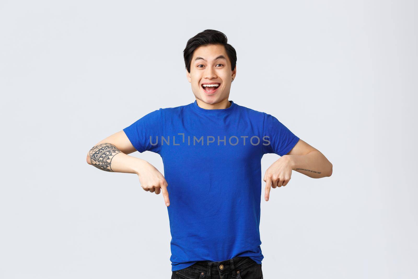 Different emotions, people lifestyle and advertising concept. Enthusiastic handsome asian man in blue t-shirt, pointing fingers down and smiling as announce good news, grey background.