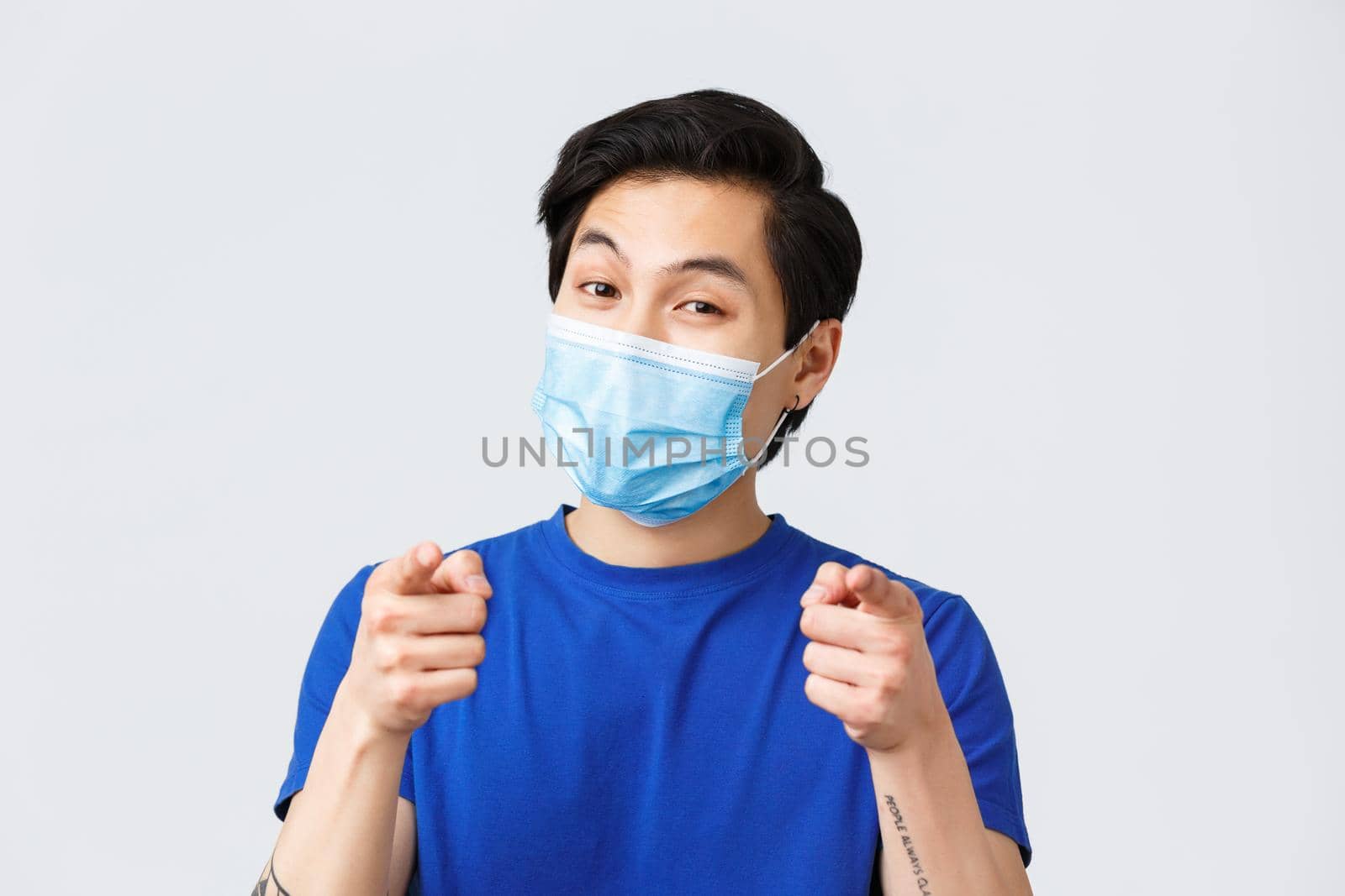 Different emotions, lifestyle and leisure during coronavirus, covid-19 concept. Close-up of attractive asian male in medical mask, guy smiling and pointing at camera, choosing.