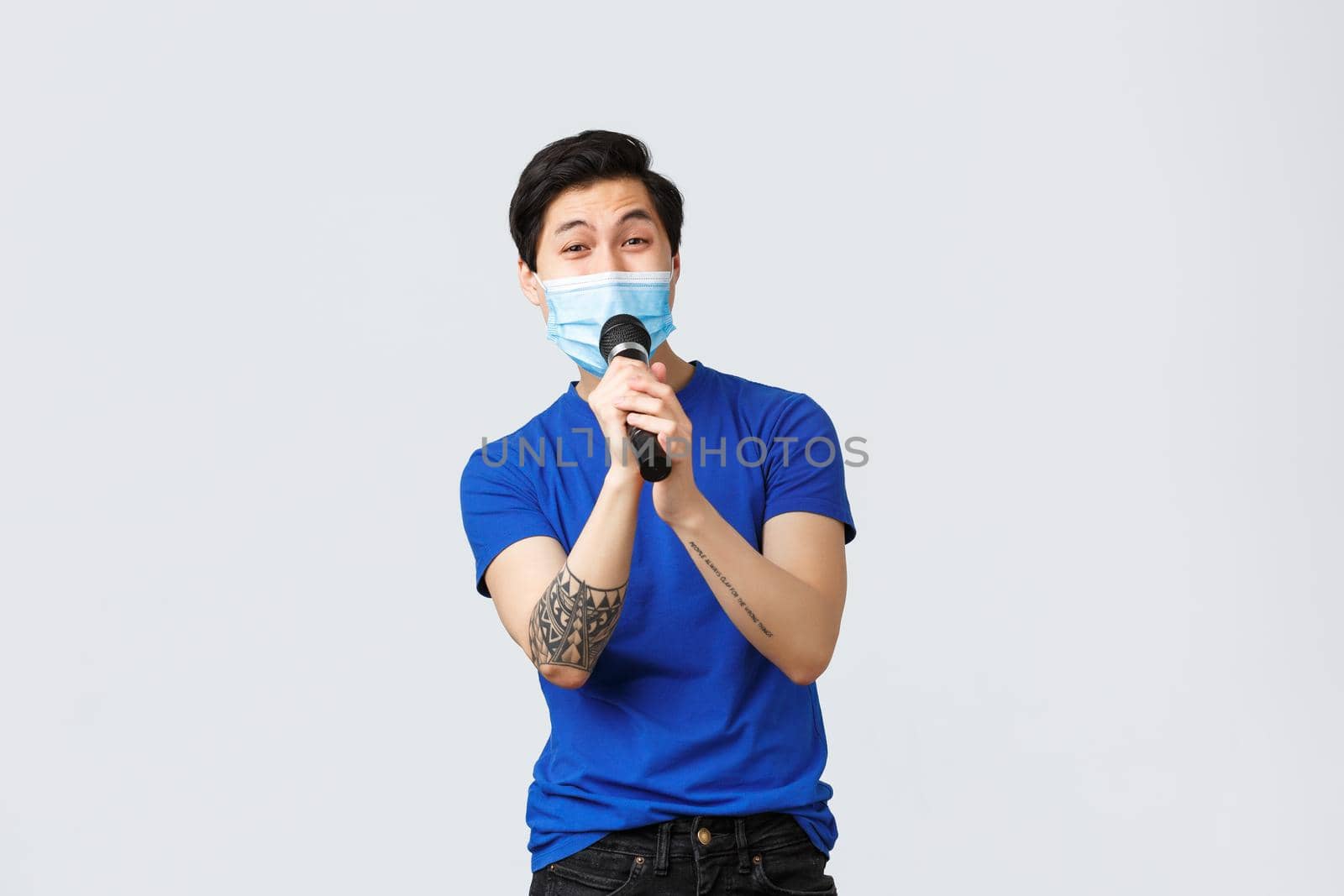 Covid-19 lifestyle, people emotions and leisure on quarantine concept. Handsome happy and carefree chinese guy having fun at karaoke, singing song in microphone, wearing medical mask.