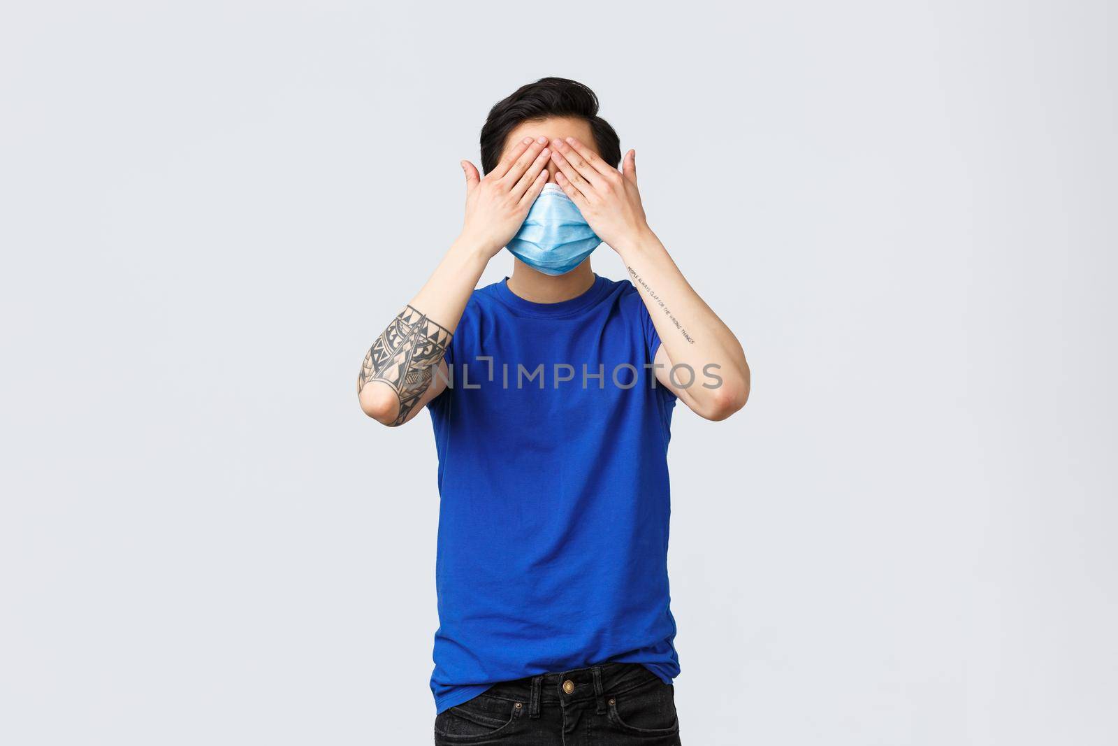 Different emotions, social distancing, self-quarantine on covid-19 and lifestyle concept. Asian man in blue t-shirt and medical mask standing blindfolded, cover eyes not see what happening by Benzoix