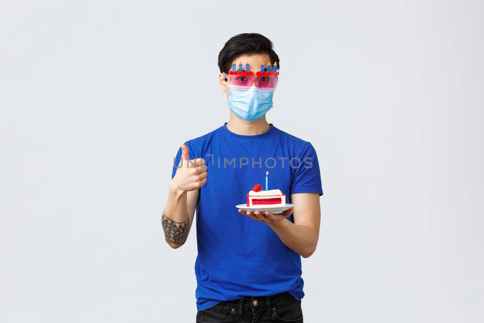 Different emotions, social distancing, self-quarantine on covid-19 and lifestyle concept. Handsome young b-day guy, asian man celebrating birthday in funny glasses, holding cake and thumb-up.