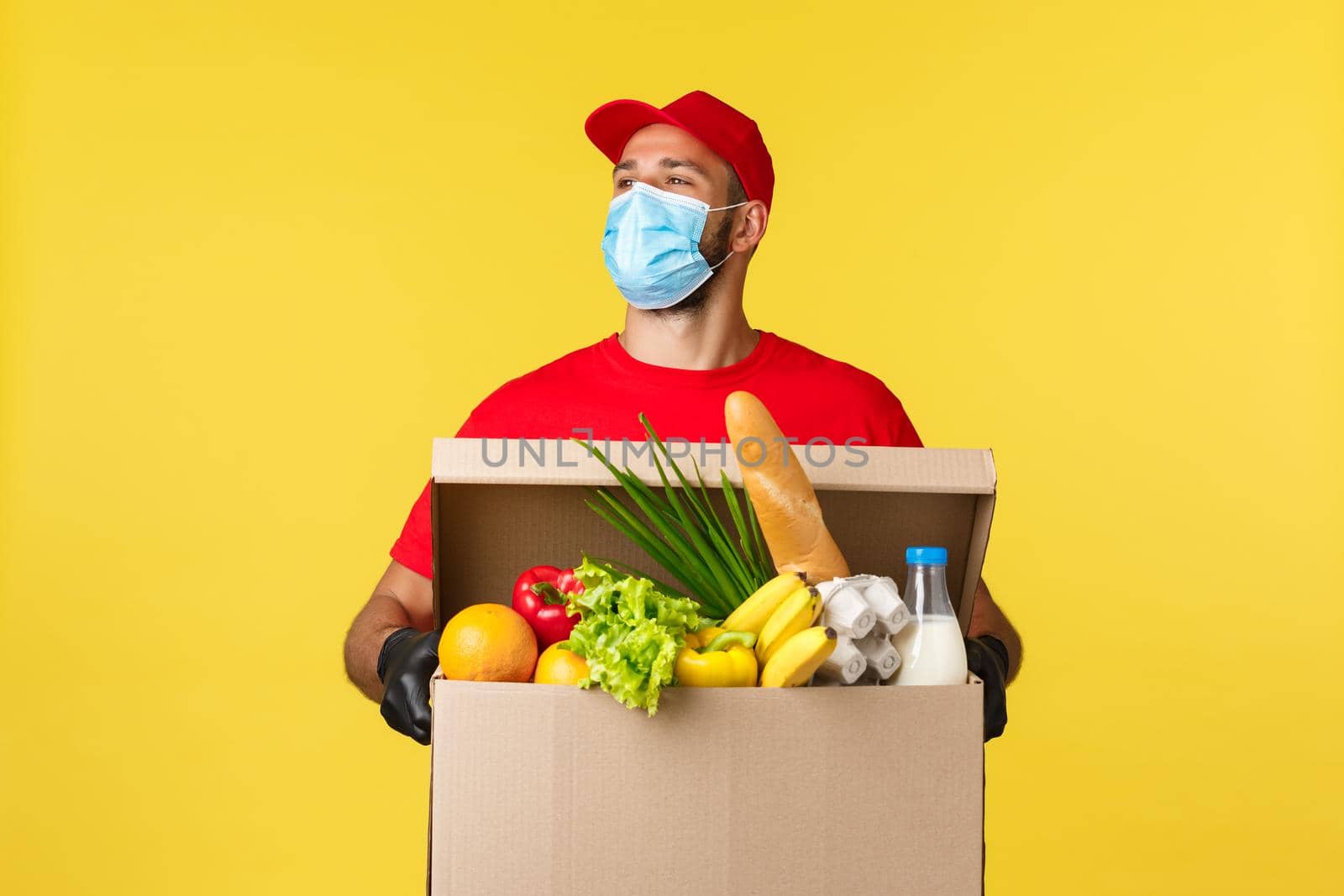 Takeaway delivery, covid-19 quarantine and groceries concept. Carefree handsome courier in red uniform and medical mask, look around with smile, holding box with food order.