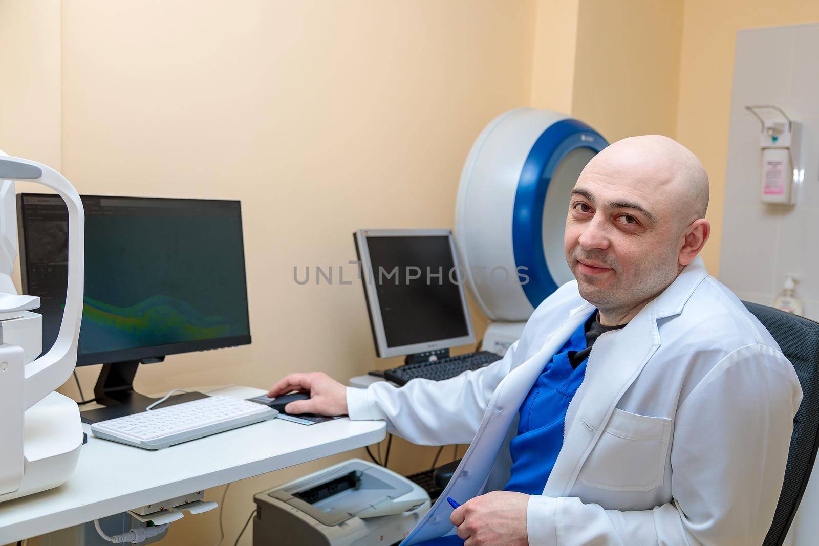 A male ophthalmologist at the workplace at the computer looks at the camera with a smile.