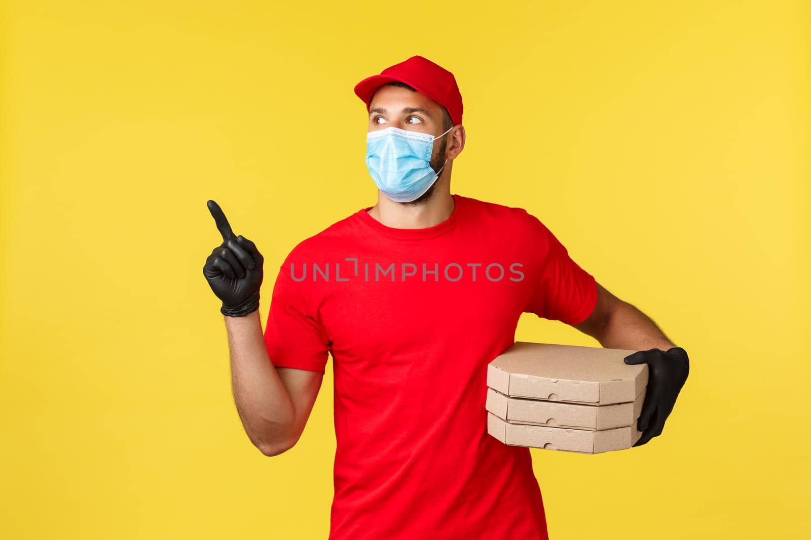 Food delivery, tracking orders, covid-19 and self-quarantine concept. Young courier searching house to deliver pizza to client, look left, wear red uniform and protective personal equipment.
