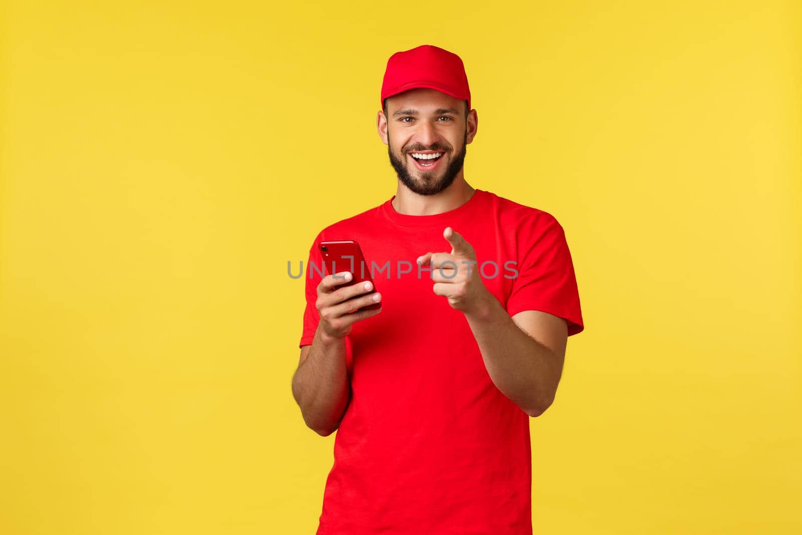 Contactless payment, online delivery, internet shopping and parcel tracking concept. Handsome bearded employee, courier guy in red t-shirt and cap, pointing at camera gotcha as using mobile phone.