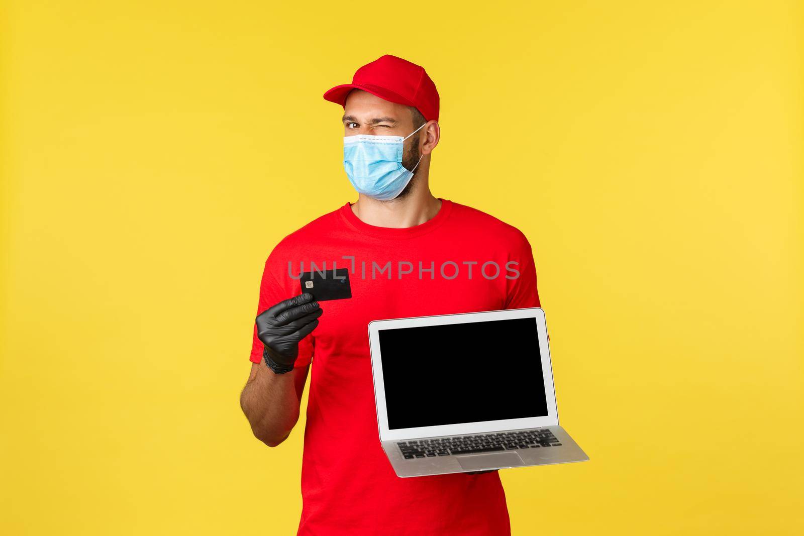 covid-19, safe shopping, express delivery and order tracking concept. Cheerful young male courier in red uniform and medical mask, recommend use contactless payment, show credit card and laptop.