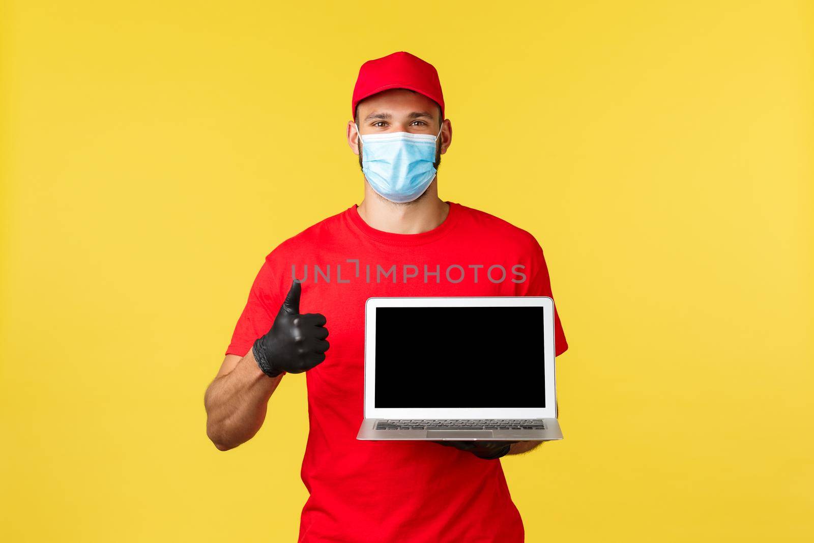 covid-19, safe shopping, express delivery and order tracking concept. Friendly young courier in red uniform and medical mask, show thumb-up, recommend visit link or site, show laptop screen.