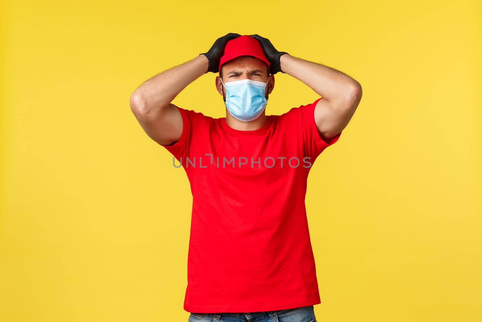 Express delivery during pandemic, covid-19, safe shipping, online shopping concept. Troubled and nervous courier in red uniform, medical mask, grab head have problem, yellow background.