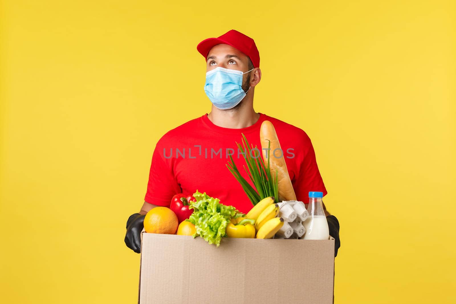 Takeaway delivery, food and drinks, covid-19 quarantine and groceries concept. Courier looking up at building, bring order grocery box to home or office, wearing protective medical mask, gloves.