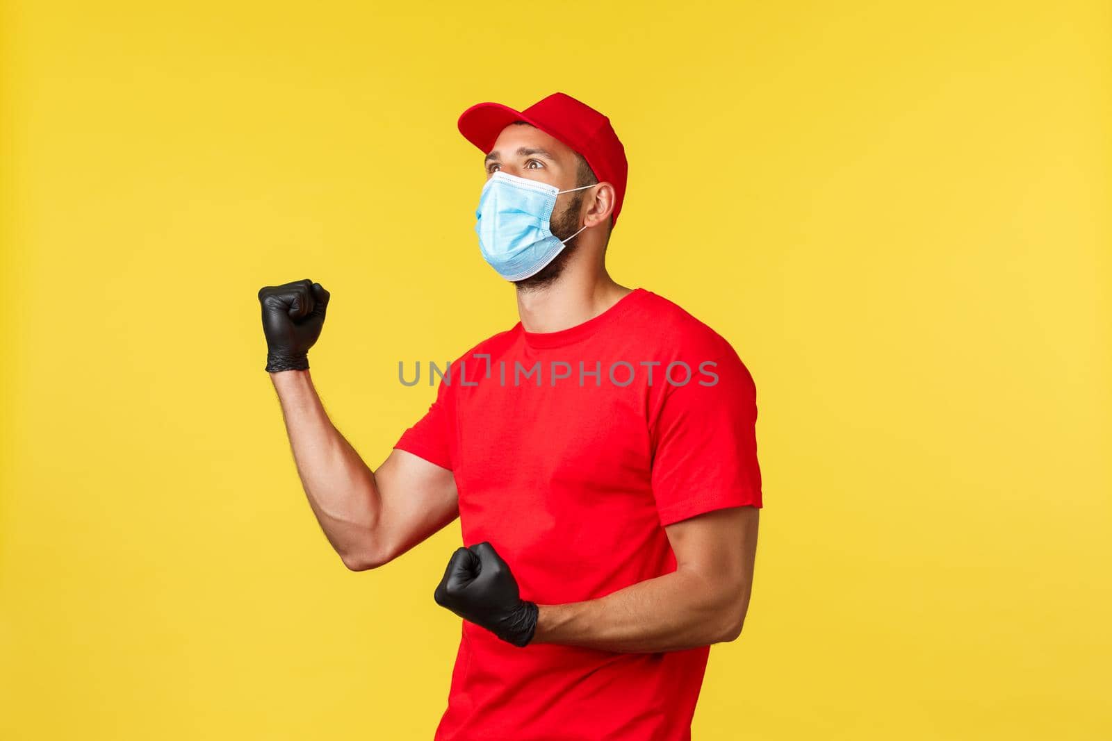 Express delivery during pandemic, covid-19, safe shipping, online shopping concept. Rejoicing happy courier in red uniform, medical mask and gloves, fist pump look upper left at screen, celebrating.