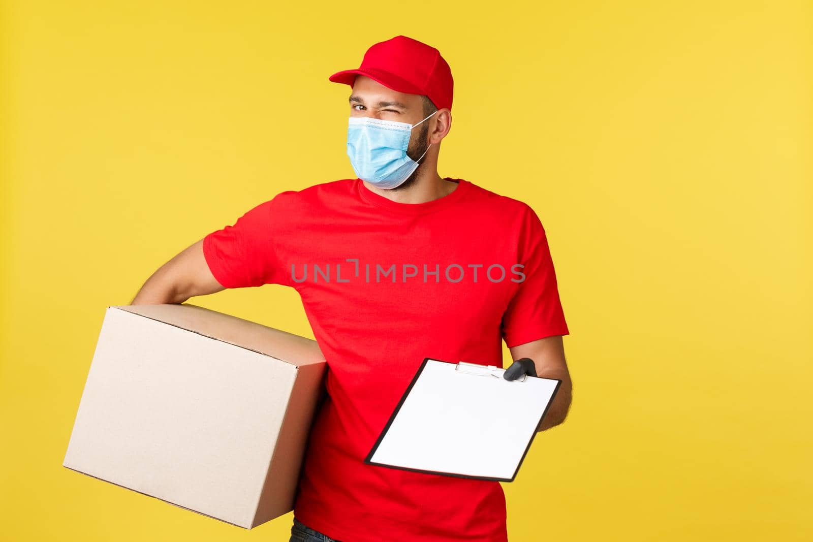 Express delivery during pandemic, covid-19, safe shipping, shopping concept. Enthusiastic handsome courier in red uniform, medical mask and gloves, hold package box and give clipboard form to sign.