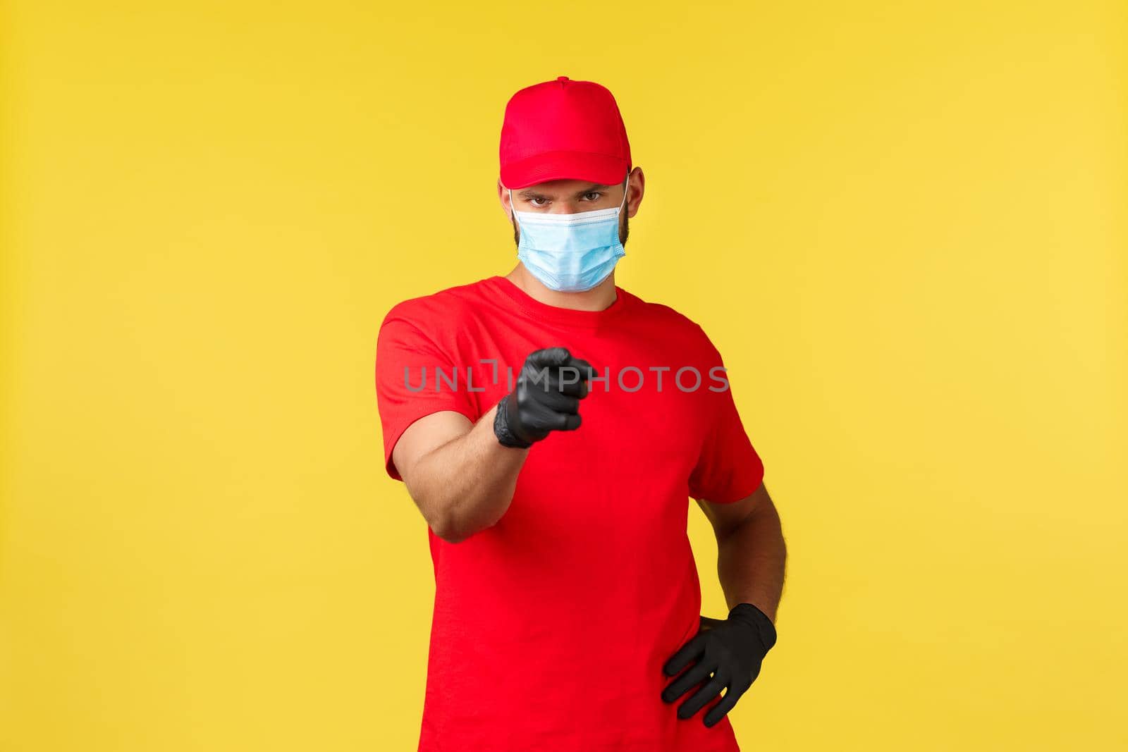 Express delivery during pandemic, covid-19, safe shipping, online shopping concept. Handsome courier in red uniform, medical mask and gloves, pointing client and make phone call sign.