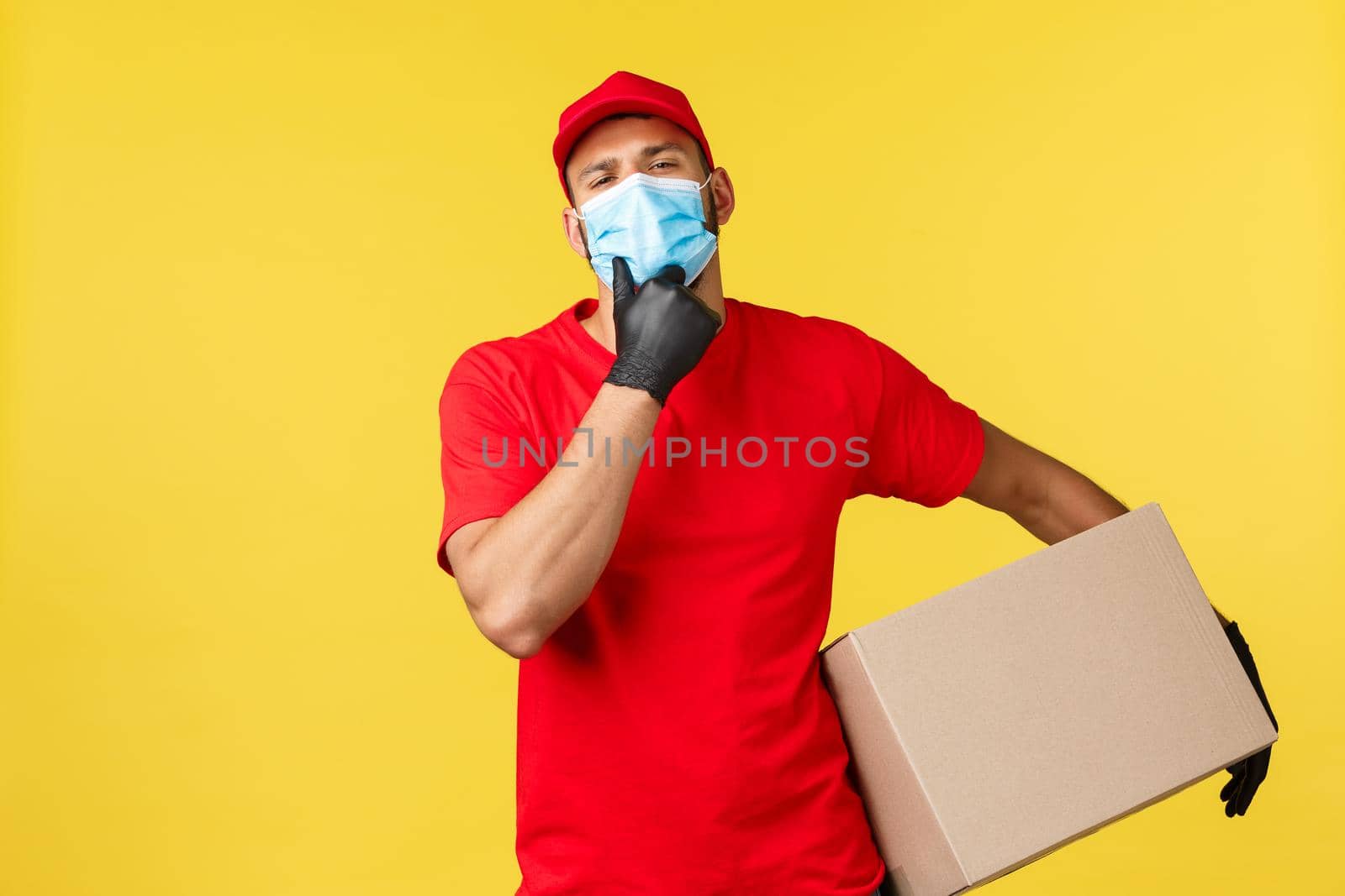 Express delivery during pandemic, covid-19, safe shipping, online shopping concept. Handsome courier in red uniform and medical mask, holding package, touch chin sassy, yellow background.