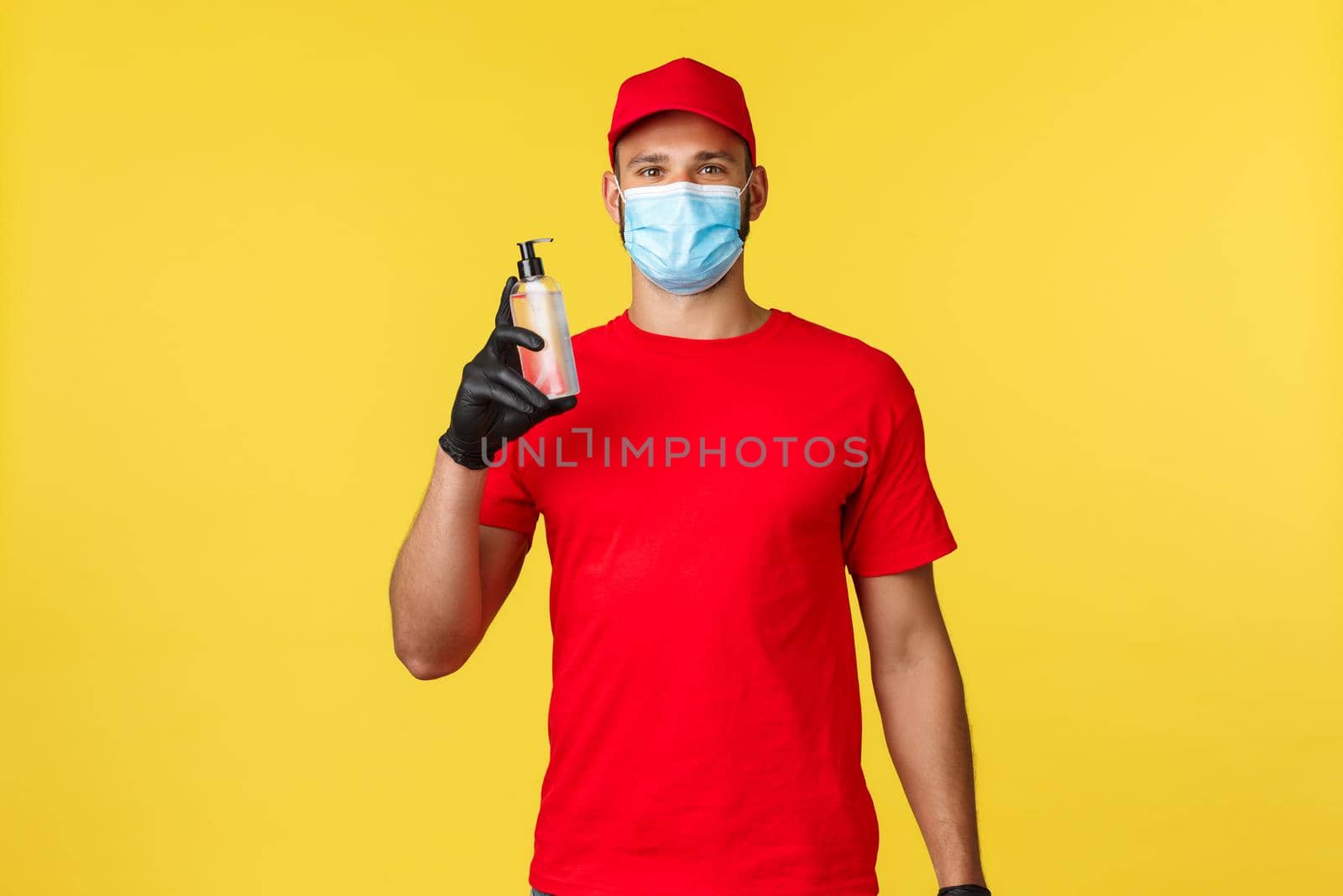 Covid-19, delivery orders, shopping, contactless handling and social distancing concept. Enthusiastic courier in red uniform, medical mask and gloves, recommend use hand sanitizer, smiling.