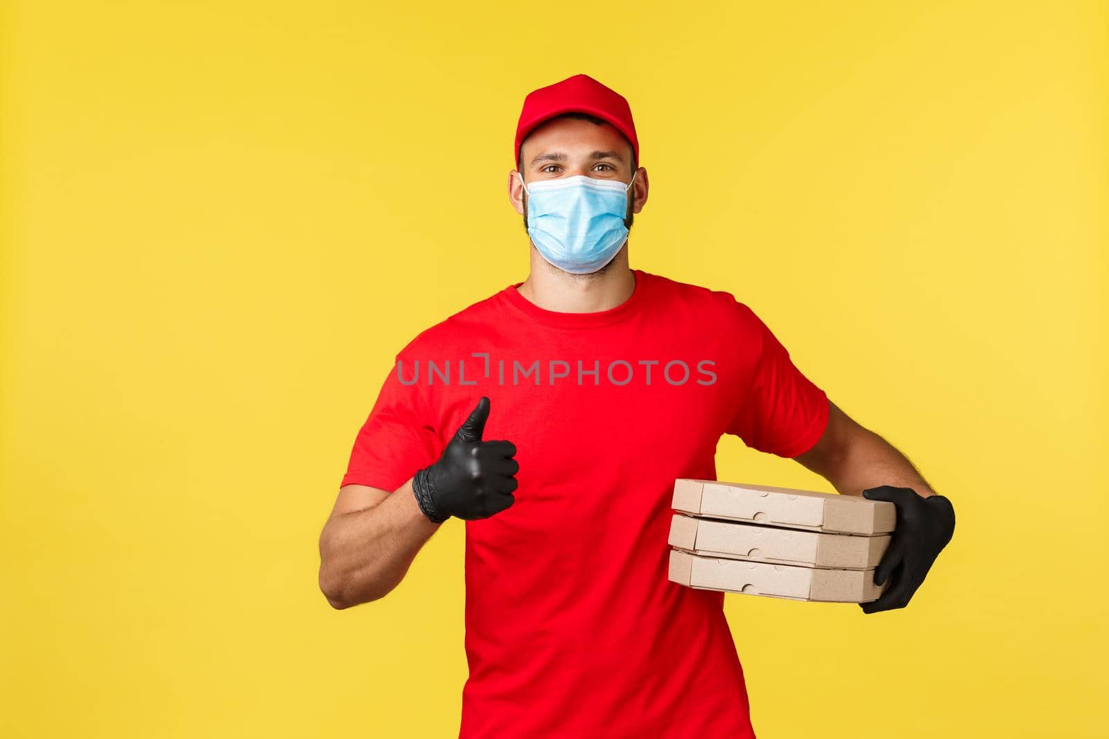 Express delivery during pandemic, covid-19, safe shipping, shopping concept. Friendly young courier in red uniform and medical mask, provide safe order delivery, holding pizzas and show thumb-up.