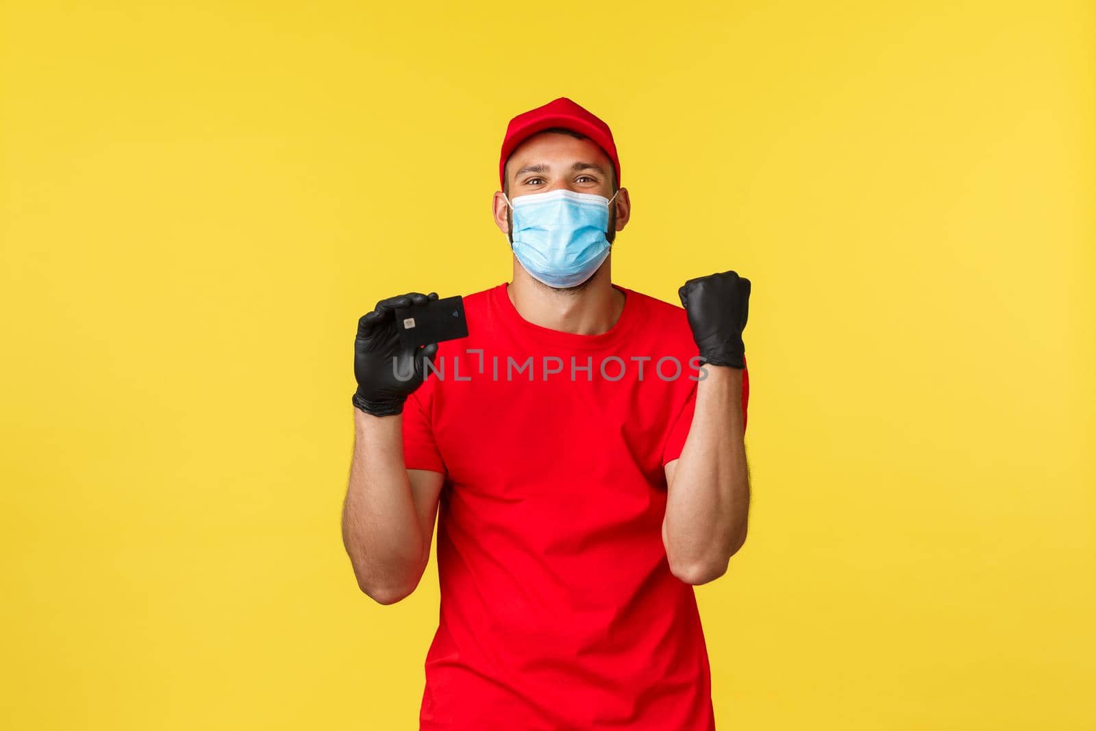 Express delivery during pandemic, covid-19, safe shipping, online shopping concept. Satisfied, happy courier in medical mask and gloves, company red uniform, chanting, say yes as showing credit card.