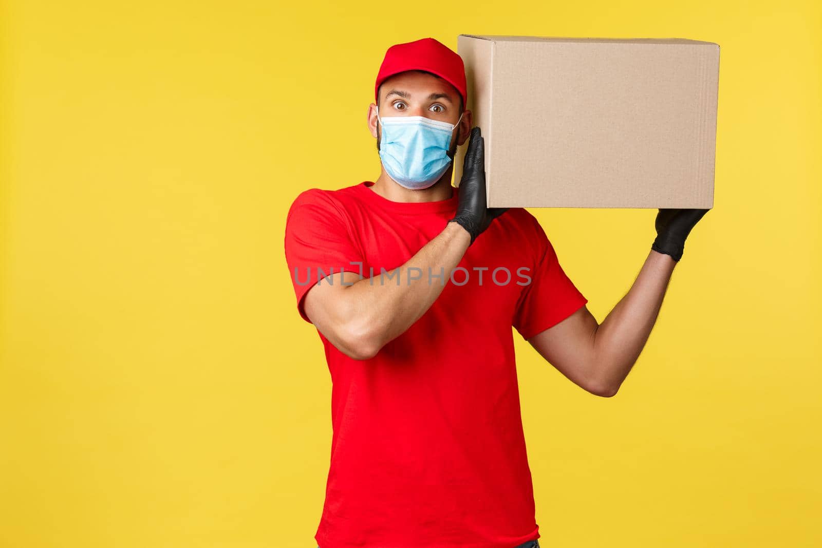 Express delivery during pandemic, covid-19, safe shipping, online shopping concept. Surprised courier in red uniform, medical mask, look excited camera, holding box package on shoulder.