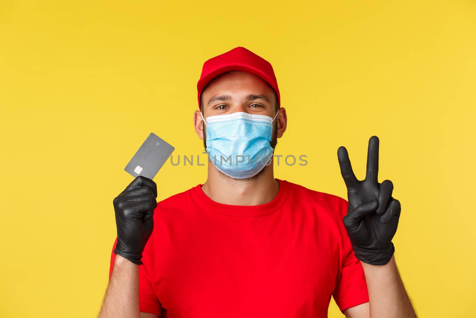 Covid-19, delivery orders, shopping, contactless payment and social distancing concept. Friendly courier, male employee in red uniform and medical mask, show peace sign and credit card.
