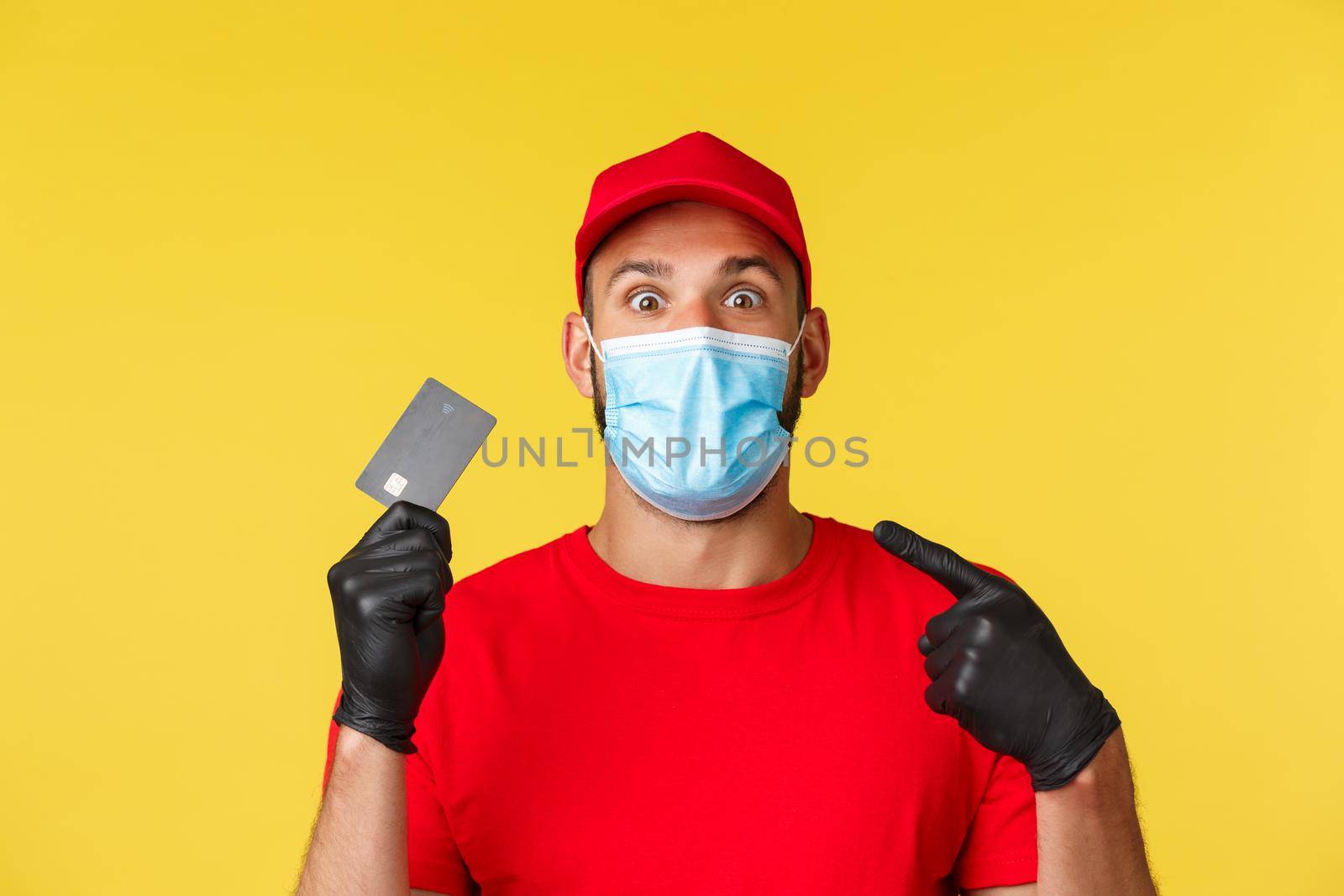 Covid-19, delivery orders, shopping, contactless payment and social distancing concept. Excited and impressed courier in red uniform and medical mask inform about credit card features.