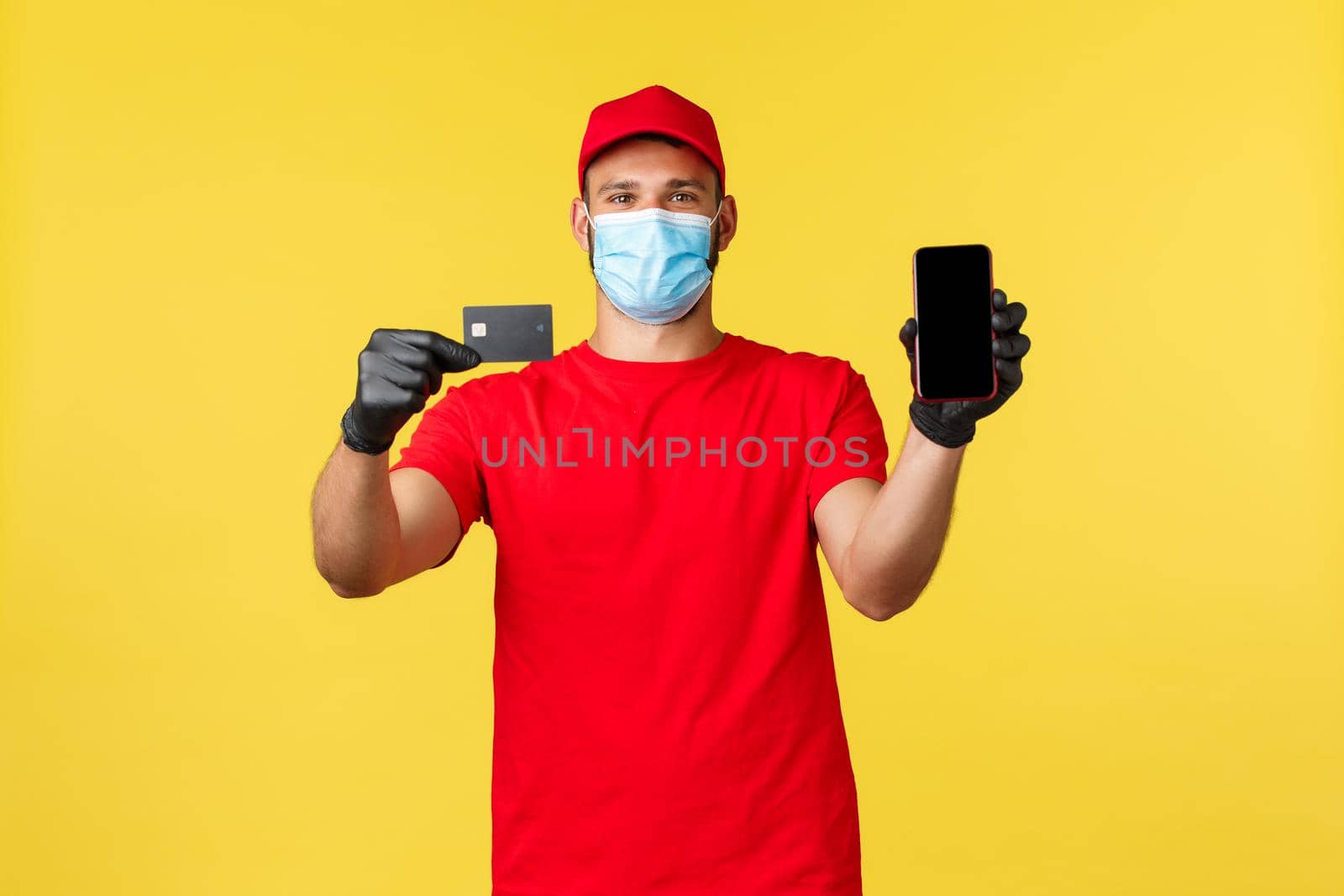 Food delivery, tracking orders, covid-19 and self-quarantine concept. Smiling courier in medical mask and gloves, showing credit card and smartphone, ways of paying for groceries deliver.