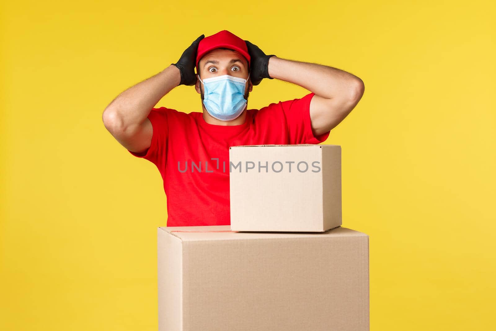 Express delivery during pandemic, covid-19, safe shipping, online shopping concept. Embarrassed and nervous, panicking courier standing near boxes with packages, gasping grab head, wear medical mask.