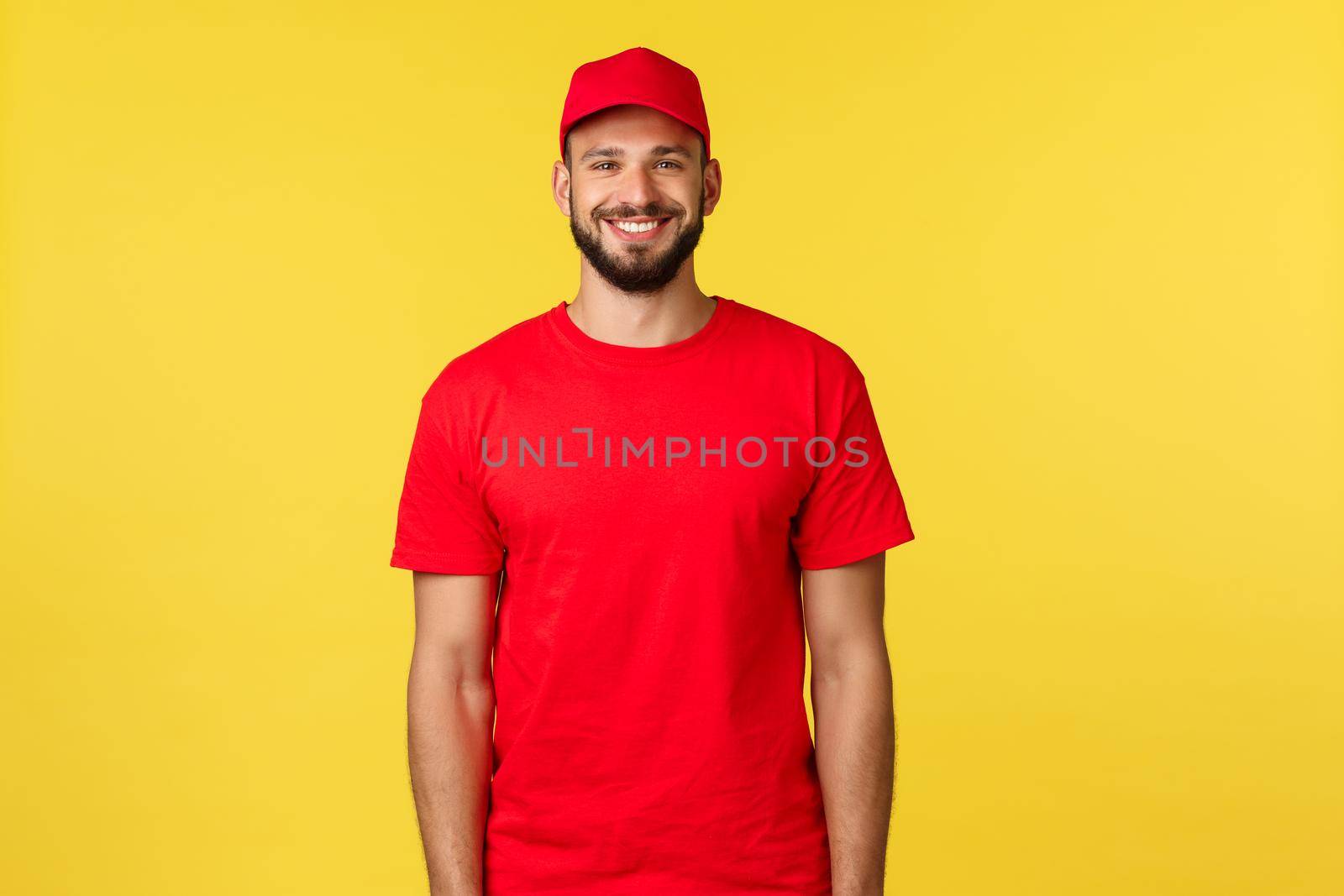 Express mail delivery, package transfer and logistics concept. Handsome friendly-looking courier in red uniform of company, t-shirt and cap, smiling broadly as standing yellow background.