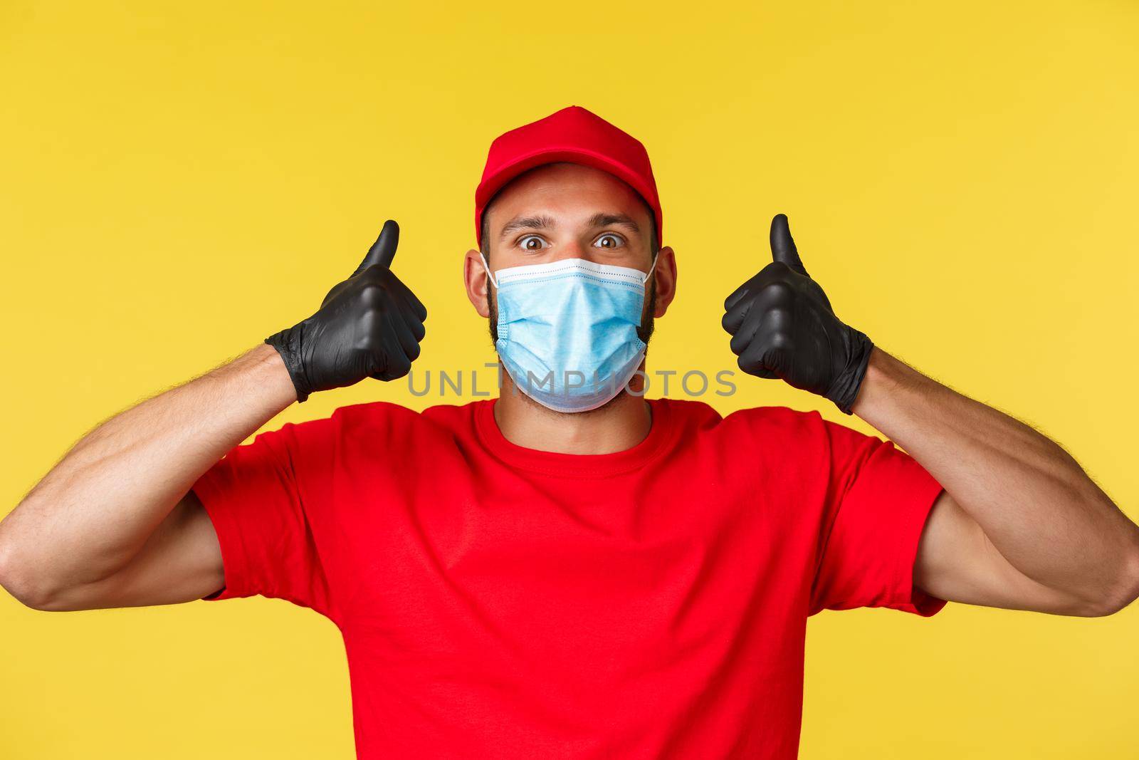 Covid-19, delivery orders, shopping, contactless payment and social distancing concept. Upbeat and excited male courier in red uniform, medical mask, show thumbs-up in approval.