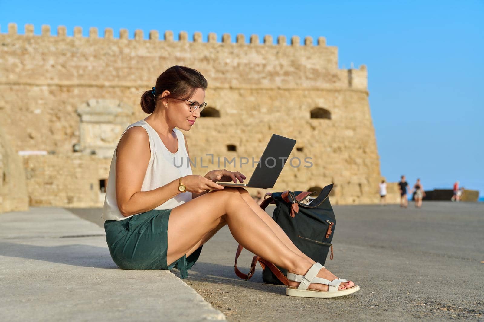 Mature woman tourist using laptop, old european historic fortress background. Technology, vacations, tourism, leisure, active lifestyle, middle aged people concept