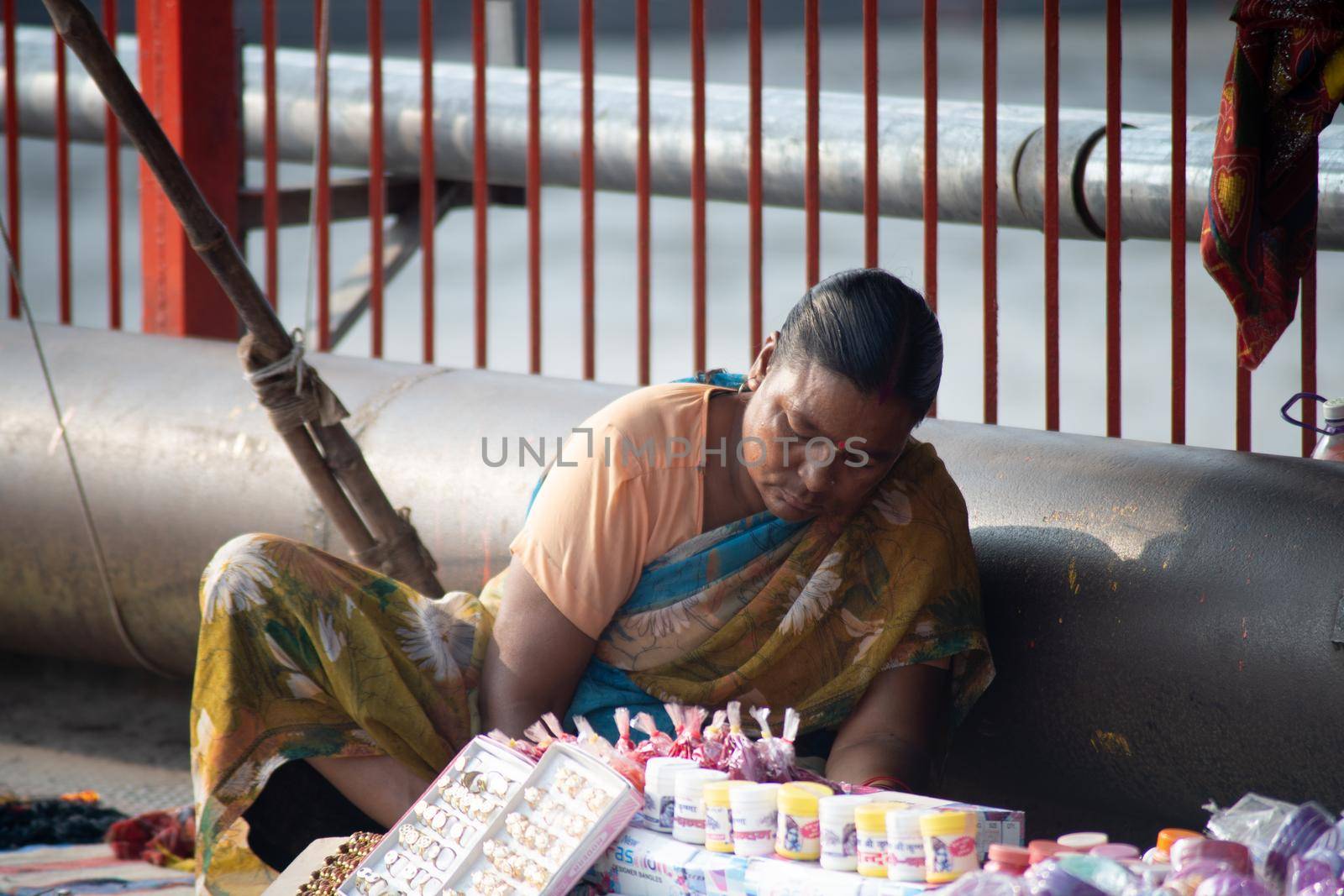 Old streetside shopkeeper making handmade bead necklaces for sale sitting under a plastic sheet, haridwar houses a lot of these old destitute people working to make ends meet by Shalinimathur