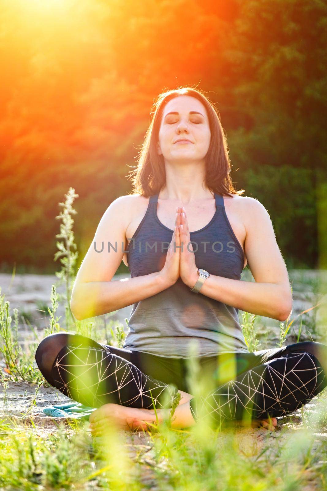 a woman practices yoga and meditates in the lotus position on the sand against the backdrop of trees. vertical
