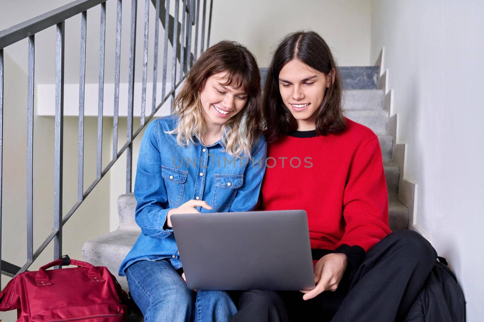 Teenage couple of friends students looking at laptop together. Teenagers male and female with backpacks sitting on the stairs inside the building. Youth, lifestyle, technology, young people concept