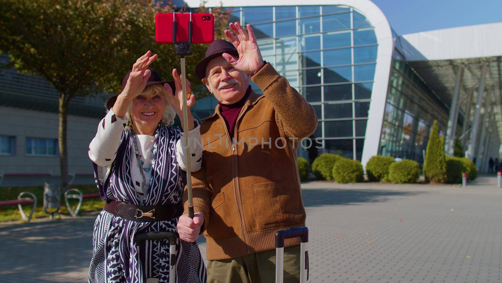 Senior couple pensioner tourists grandmother grandfather with luggage suitcases using mobile phone make selfie vlog in social media near airport hall, railway station. Travel, vacation, journey, trip