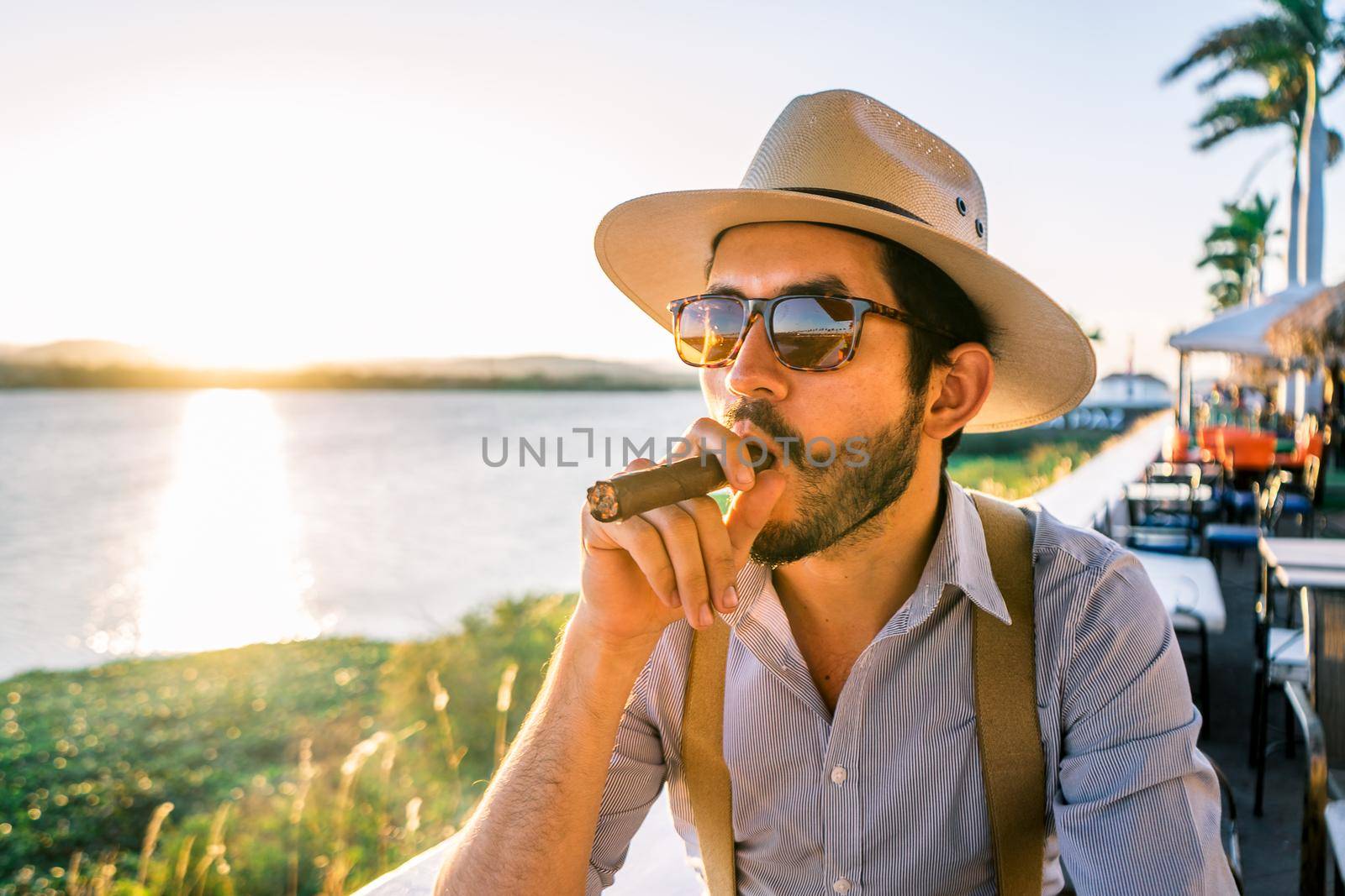 Latin hipster in a bar on the shore of a lake at sunset wearing a hat and suspenders and smoking a cigar by cfalvarez