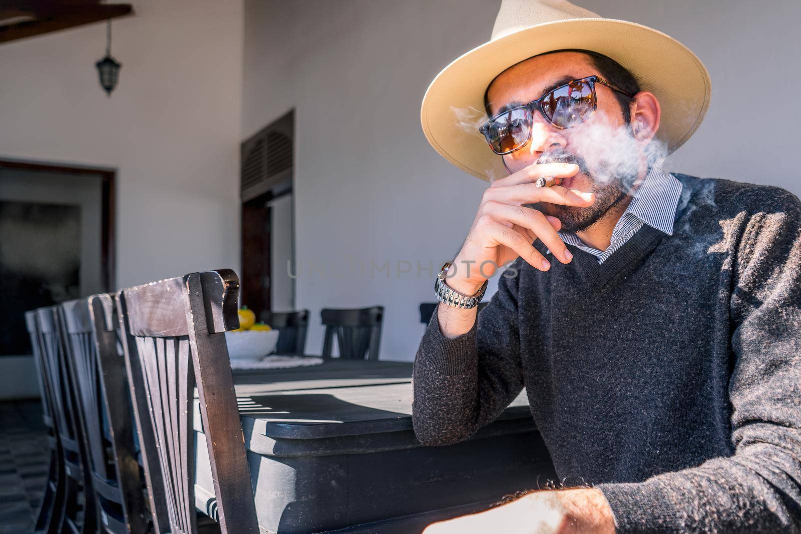 Young Nicaraguan man wearing a sweater hat and sunglasses smoking a cigar in the living room of a colonial-style mansion in Managua by cfalvarez