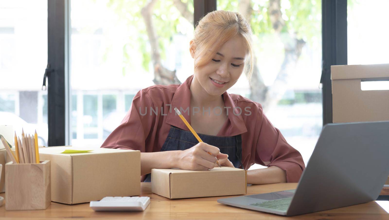 Close up woman sitting in the office full of packages in the background emplaning on a laptop and using a calculator, for online business, SME, finance, e-commerce and delivery concept..